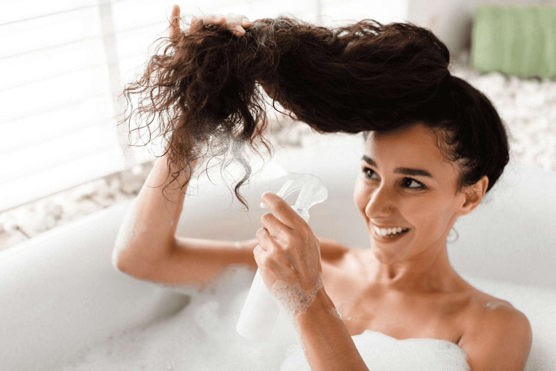 A young woman applies essential oil spray on her hair while lying in a bubble bath at home.